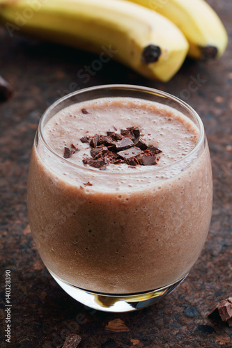 Close-up of a whipped cold banana milkshake in a glass with chocolate chips on a dark brown table and branches of bananas