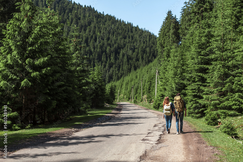 The couple go with travel backpacks on the edge of the road near a pine forest. Active holidays in the mountains