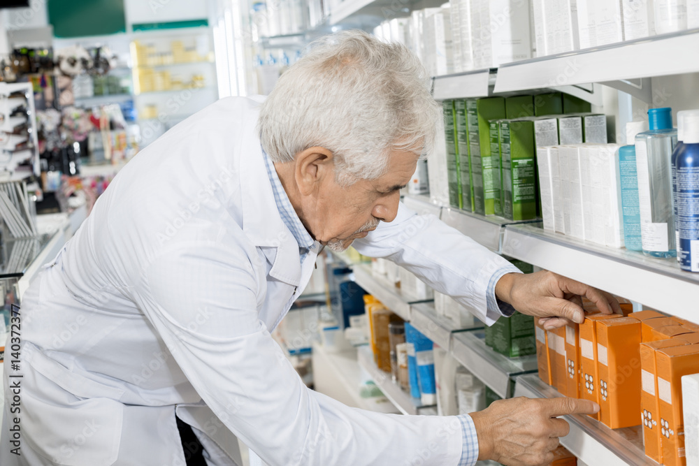 Male Pharmacist Arranging Medicines On Shelf In Pharmacy Male Pharmacist Arranging Medicines On Shelf In Pharmacy