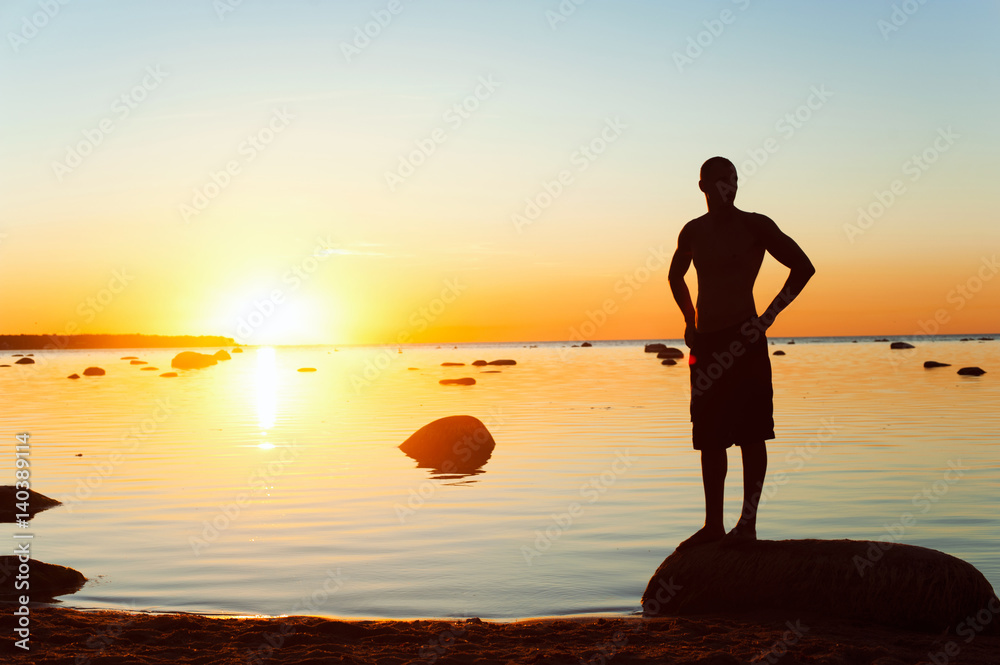 Young sporty man silhouette standing on the seashore at sunset
