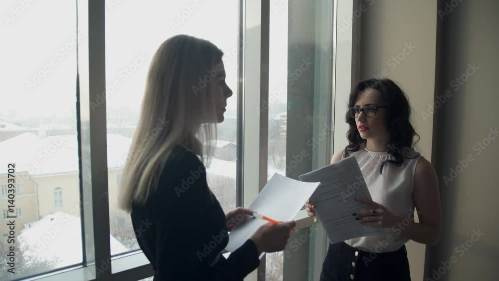 Two business women against window in hall discuss topics. tall blonde woman with straight hair holding folder with paper, pen, listening to employee, says, writes important points in synopsis in hall