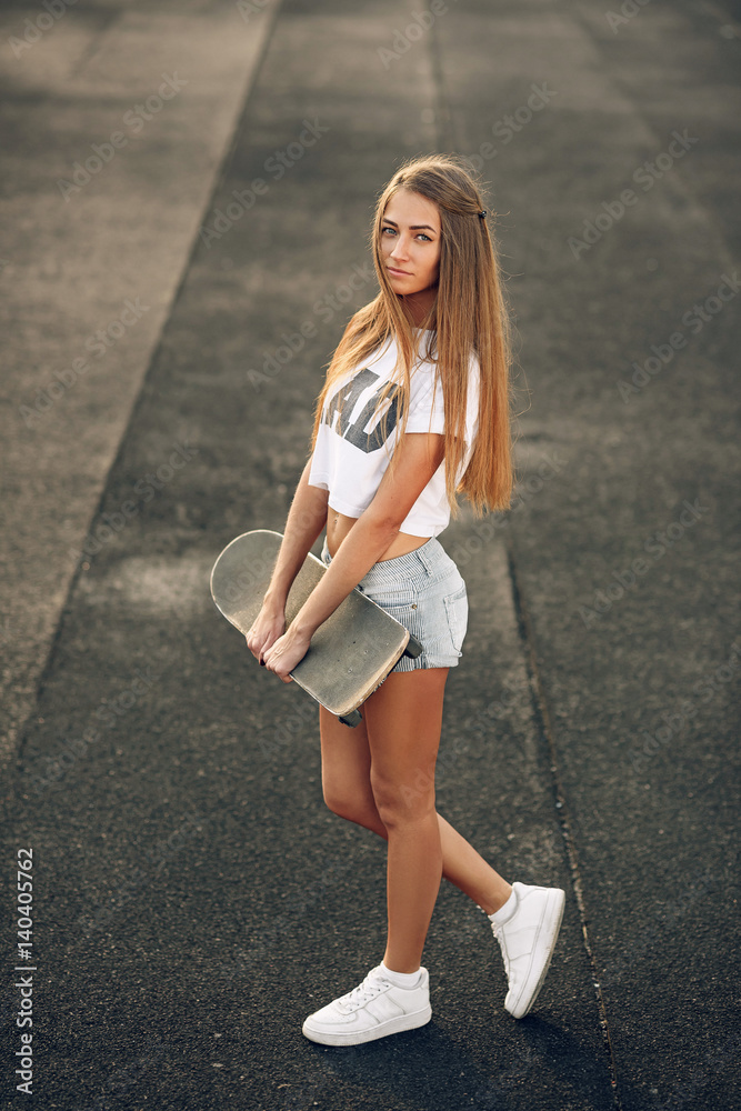 Beautiful and sexy young girl in a white T-shirt and shorts standing with skateboard in hands