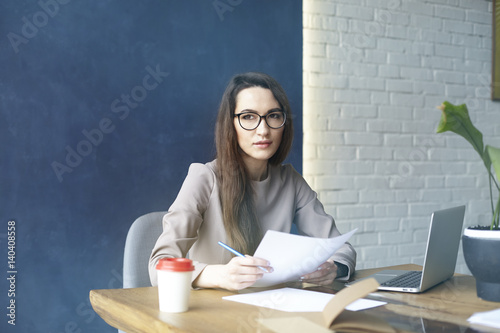 Beautiful businesswoman with long hair reading a book while sitting in his modern loft office. Dark blue wall background, day light