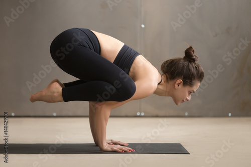 Young attractive smiling yogi woman practicing yoga, standing in Bakasana exercise, Crane pose, working out, wearing black sportswear, cool urban style, full length, grey studio background, side view