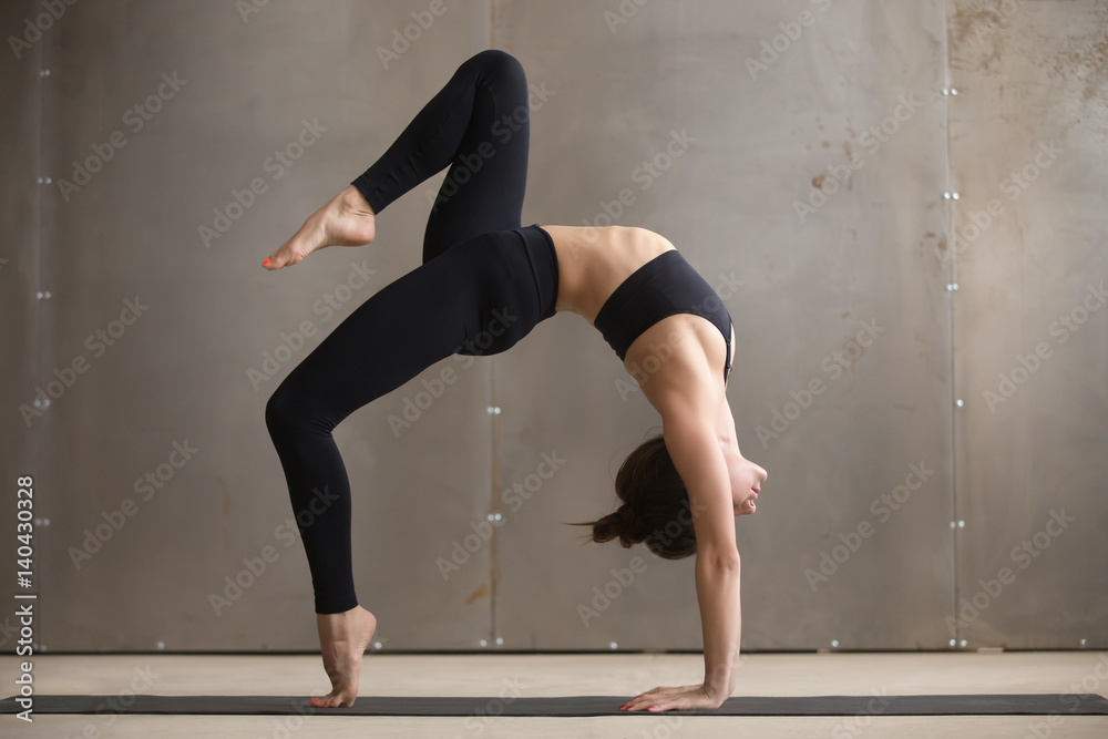 Young attractive yogi woman practicing yoga, standing in Bridge exercise, One legged Wheel pose, working out, wearing black sportswear, cool urban style, full length, grey studio background, side view