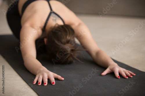 Young attractive woman practicing yoga, stretching in Child exercise, Balasana pose, working out, wearing black sportswear, urban style grey studio floor, close up, focus on fingers