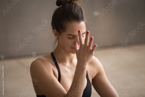 Young attractive yogi woman practicing yoga, using Alternate Nostril Breathing, making nadi shodhana pranayama therapeutic exercise, working out, cool urban style grey studio background, closeup, photo