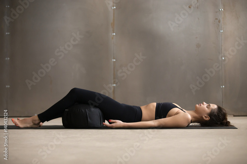 Young attractive sporty yogi woman practicing yoga, lying in Savasana exercise, Dead Body, Corpse pose using Zafu cushion for comfort, resting after working out, urban style grey studio, full length