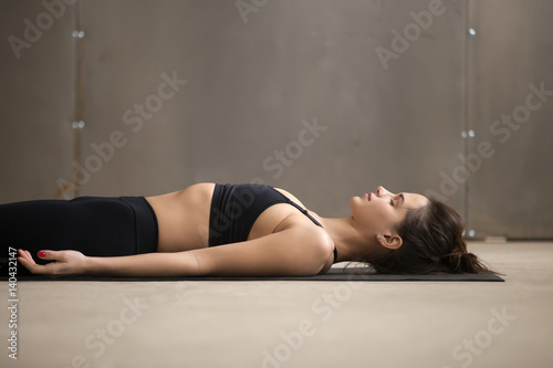 Young attractive woman practicing yoga, lying in Dead Body, Savasana exercise, Corpse pose, working out, wearing black sportswear, cool urban style, closeup, grey studio background