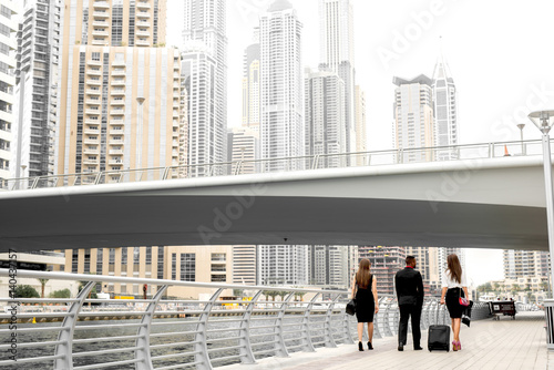 Back view of business man and two businesswomen carrying luggage walking down Downtown under bridge.