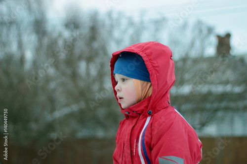 Pensive little boy sits on top of a snowy hill in bright clothes. On the street, in the village.