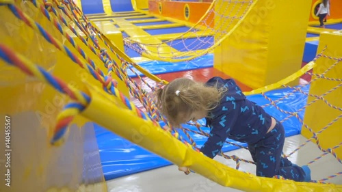 Girl is Walking by the Bridge Through rope Stairs Little Girl with Long Fair Hair in Indoors Playground. Active Toddler Girl Having Fun At Sport Center. Slow Motion photo