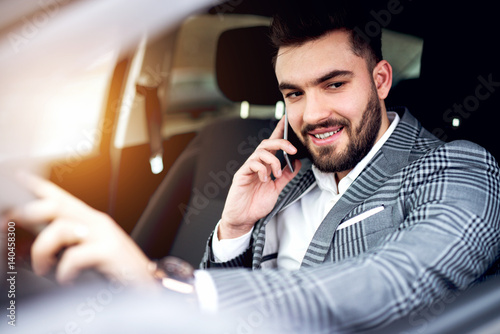 Nice bearded guy in stylish suit talking over phone smiling while driving car to meeting.