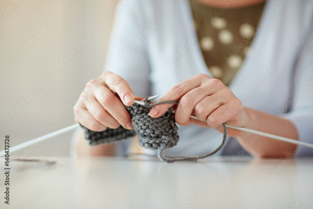 Fototapeta premium Closeup portrait of gentle hands of mature woman knitting scarf at table in home interior