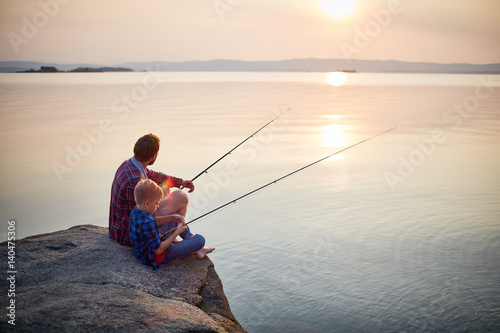 Back view portrait of father and son sitting together on rocks fishing with rods in calm lake waters with landscape of setting sun