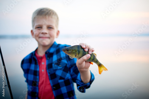 Portrait of teen blond boy smiling proudly holding one perch fish and showing it to camera against calm blue lake