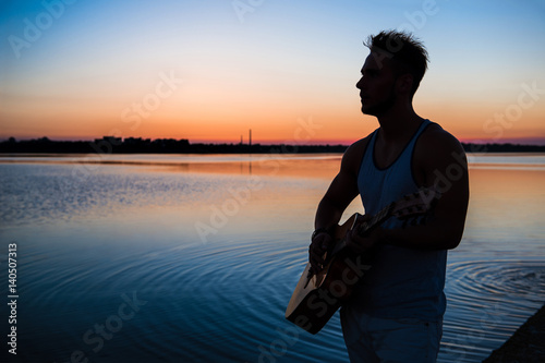 Silhouette of young handsome man playing guitar at seaside during sunrise. Outdoors.