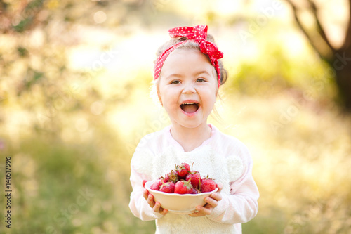Laughing baby girl 4-5 year old holding white bowl with fresh strawberries over nature green background outdoors. Looking at camera. Healthy eating. Summer time.