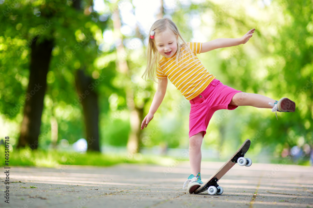 Pretty little girl learning to skateboard on beautiful summer day in a park