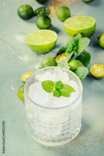 Ingredients for mojito cocktail, whole, sliced lime and mini limes, mint leaves, brown crystal sugar over gray stone texture background with glass full of ice. Space for text