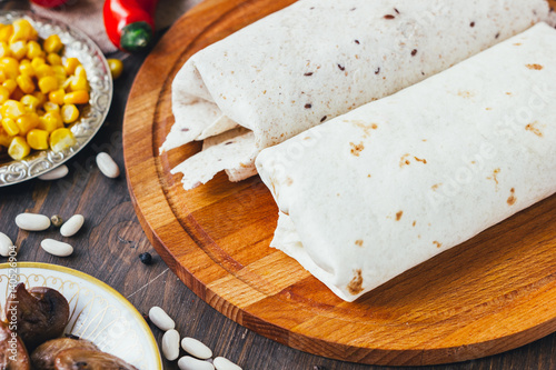 Vegetarian burrito on wooden board over black table surrounded by ingredients.
