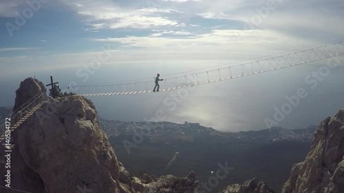 Apic aerial shot of man walking suspension bridge to Cross in Crimea. High rocks Ai-Petri of Crimean mountains. Black sea coast city and blue sky with clouds.