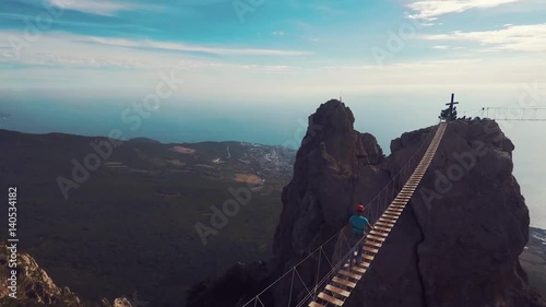 Apic aerial shot of man walking suspension bridge to Cross in Crimea. High rocks Ai-Petri of Crimean mountains. Black sea coast city and blue sky with clouds.