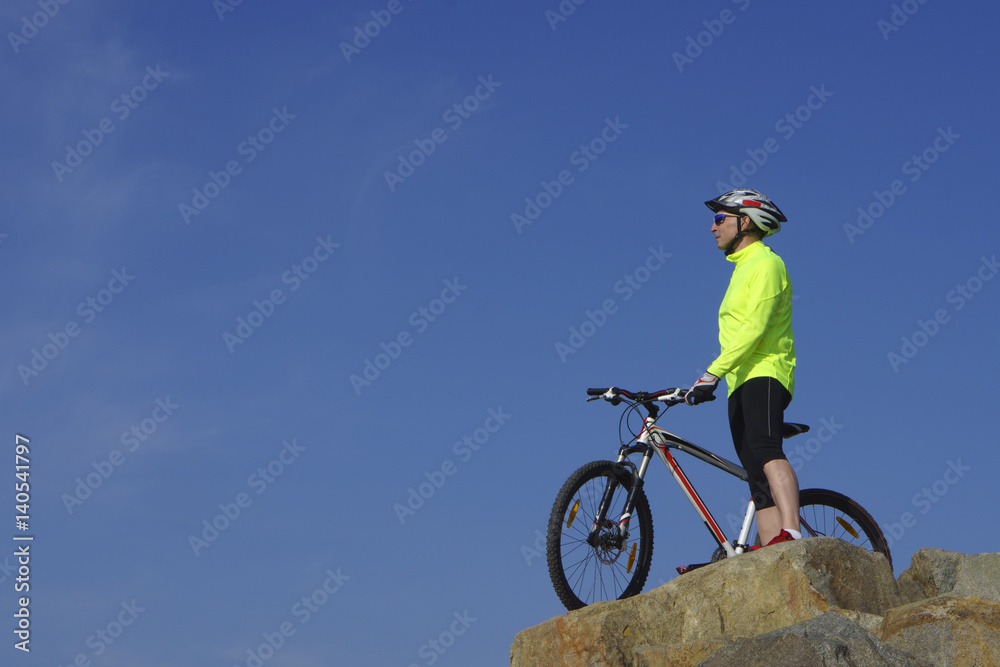 Fototapeta premium Man, in light-green jacket and red sneakers, near mountain bike with sky background