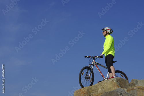 Man, in light-green jacket and red sneakers, near mountain bike with sky background