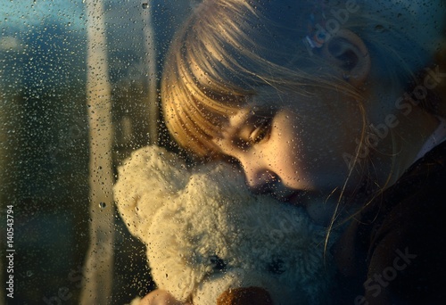 Sad child hugs a plush toy at rainy window.