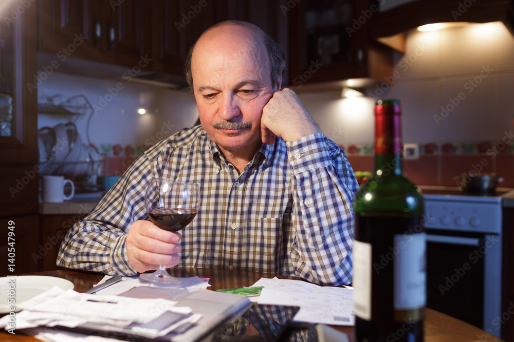 An elderly man considers a family budget and tiredly drinks red wine, trying to relax An elderly man considers a family budget and tiredly drinks red wine, trying to relax