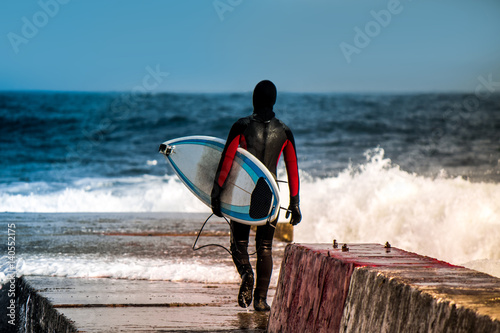 Surfer goes into water wearing a wetsuit in winter. Cold surfing. Wave splash. waterproof suit
