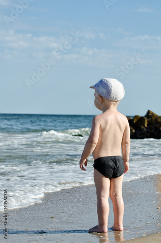 Little blond boy on the beach looking at the waves