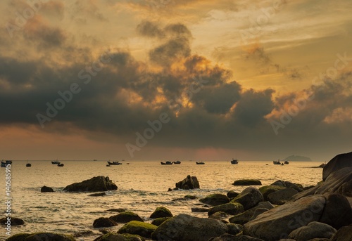 A fiery orange morning sky looking out over the south China sea in Vung Lam Bay Vietnam. With a rock covered coastline and fishing boat silhouettes.