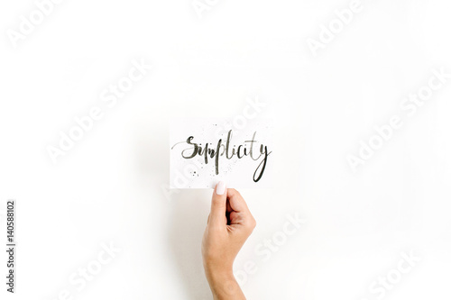 Minimal pale composition with girl's hand holding card with word Simplicity written in calligraphic style on paper on white background. Flat lay, top view