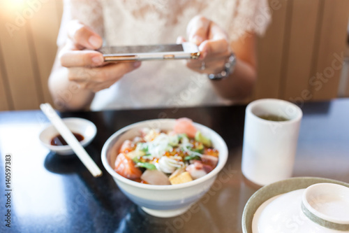 Blurred photo of young woman photographing Japanese food by her smartphone in Japanese restaurant.