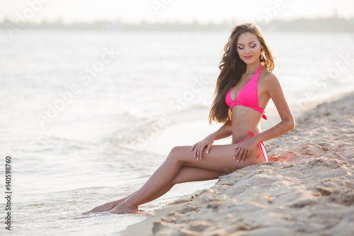 Beautiful young woman in a pink bikini sits on a beach. Horizontal shot.