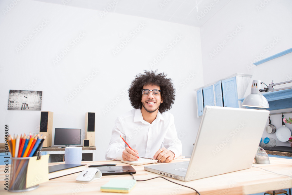 Happy hipster freelance man smiling for camera while working at table in front of laptop computer at home alone. Handsome man in glasses editing photos or videos.