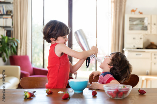 Smiling child pours whipped cream in his brother's mouth with a dispenser