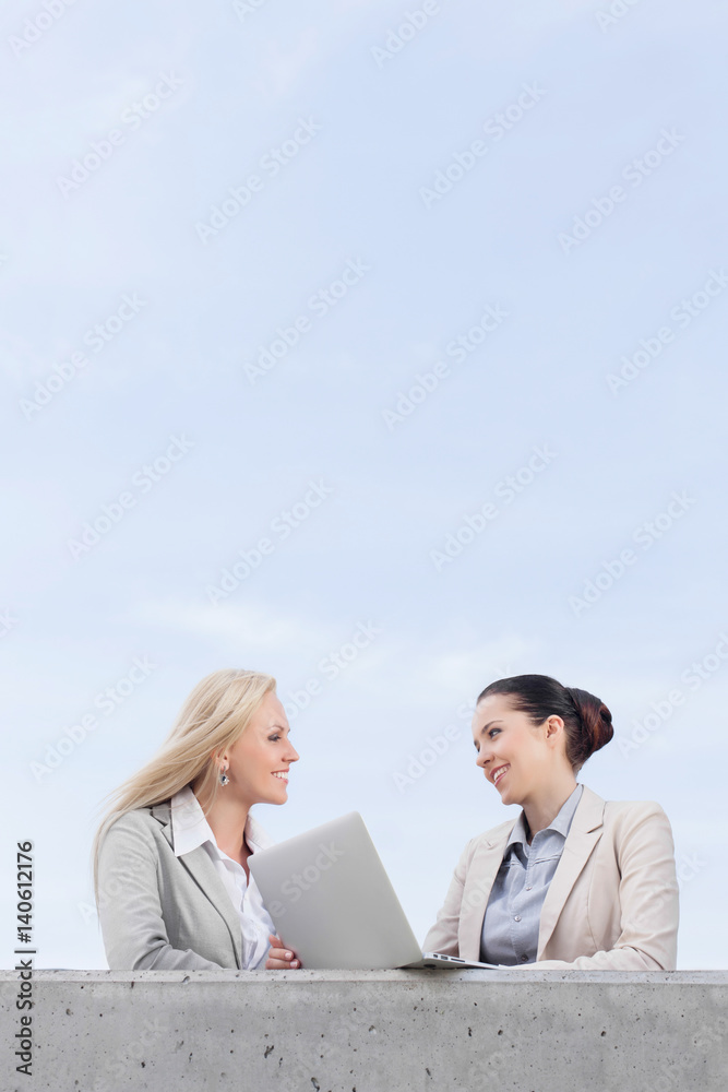 Low angle view of happy young businesswomen with laptop discussing while standing on terrace against sky