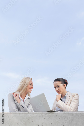 Low angle view of concentrated businesswoman using laptop while standing with coworker on terrace against sky