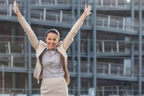 Excited young businesswoman with arms raised standing against office building