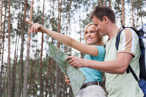 Happy young hiking couple holding map with woman pointing away in forest