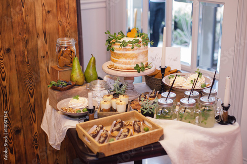 sweet bar with eclairs, lemonade,sweets and white cake decorated with greenery in area of wedding party