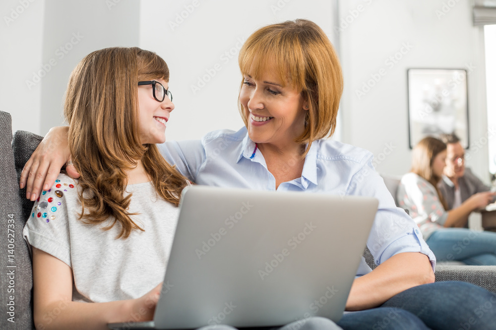 Happy mother and daughter with laptop while family sitting in background at home Happy mother and daughter with laptop while family sitting in background at home