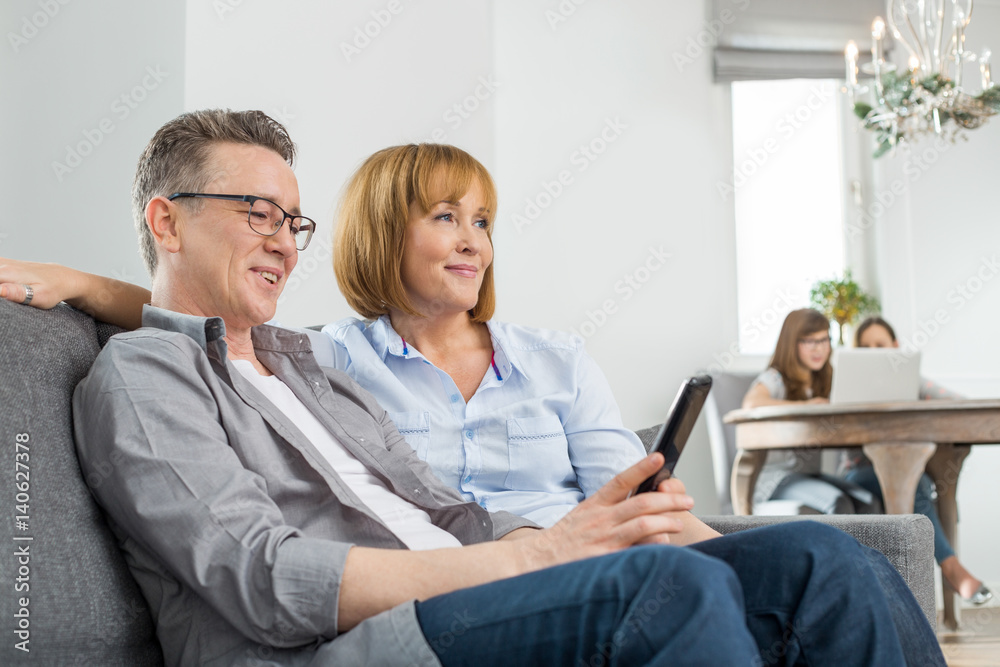 Parents watching TV on sofa with children using laptop in background Parents watching TV on sofa with children using laptop in background