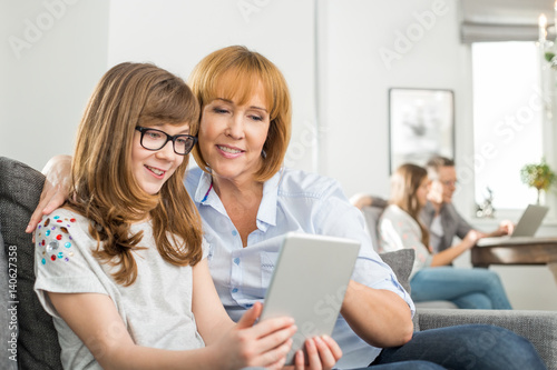 Loving mother and daughter using tablet PC with family sitting in background at home