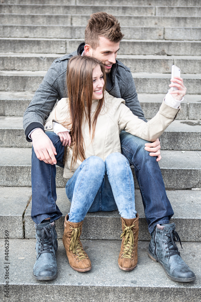 Full length of young couple taking picture of themselves while sitting on steps outdoors Full length of young couple taking picture of themselves while sitting on steps outdoors