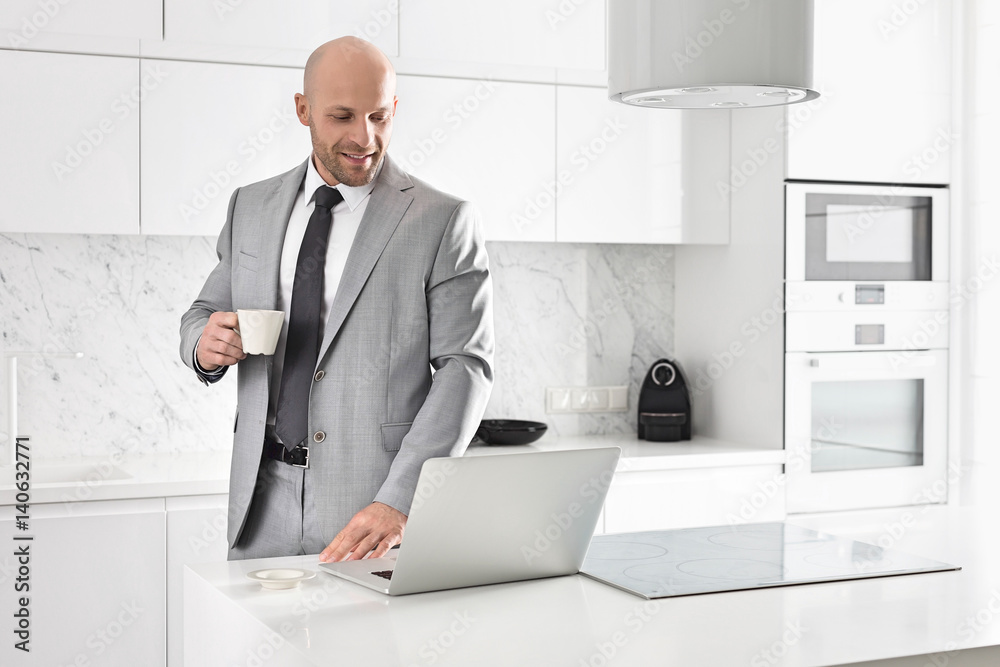 Mid adult businessman having coffee while using laptop in kitchen