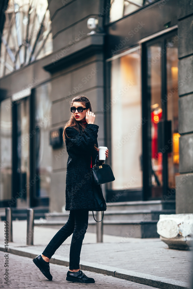 Fototapeta premium young stylish girl, passing by the Windows.Wearing fashionable glasses and a black coat.Keeps coffee.